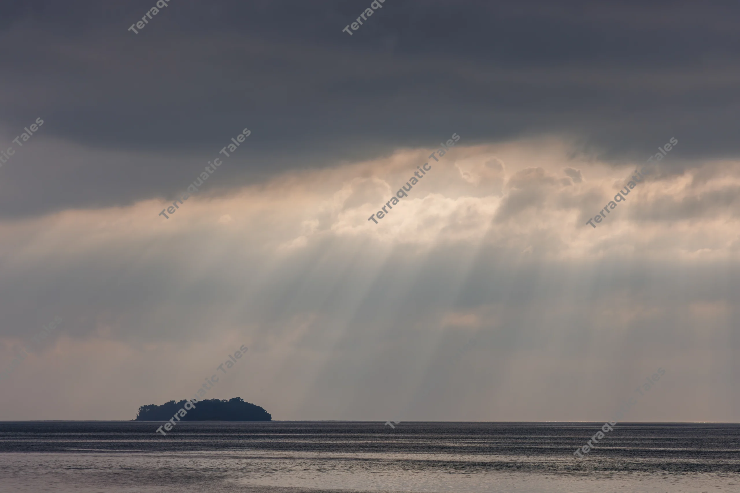 Sunrays beam through clouds above a dark, silhouetted tropical island and calm blue ocean waters in the Andaman Islands.