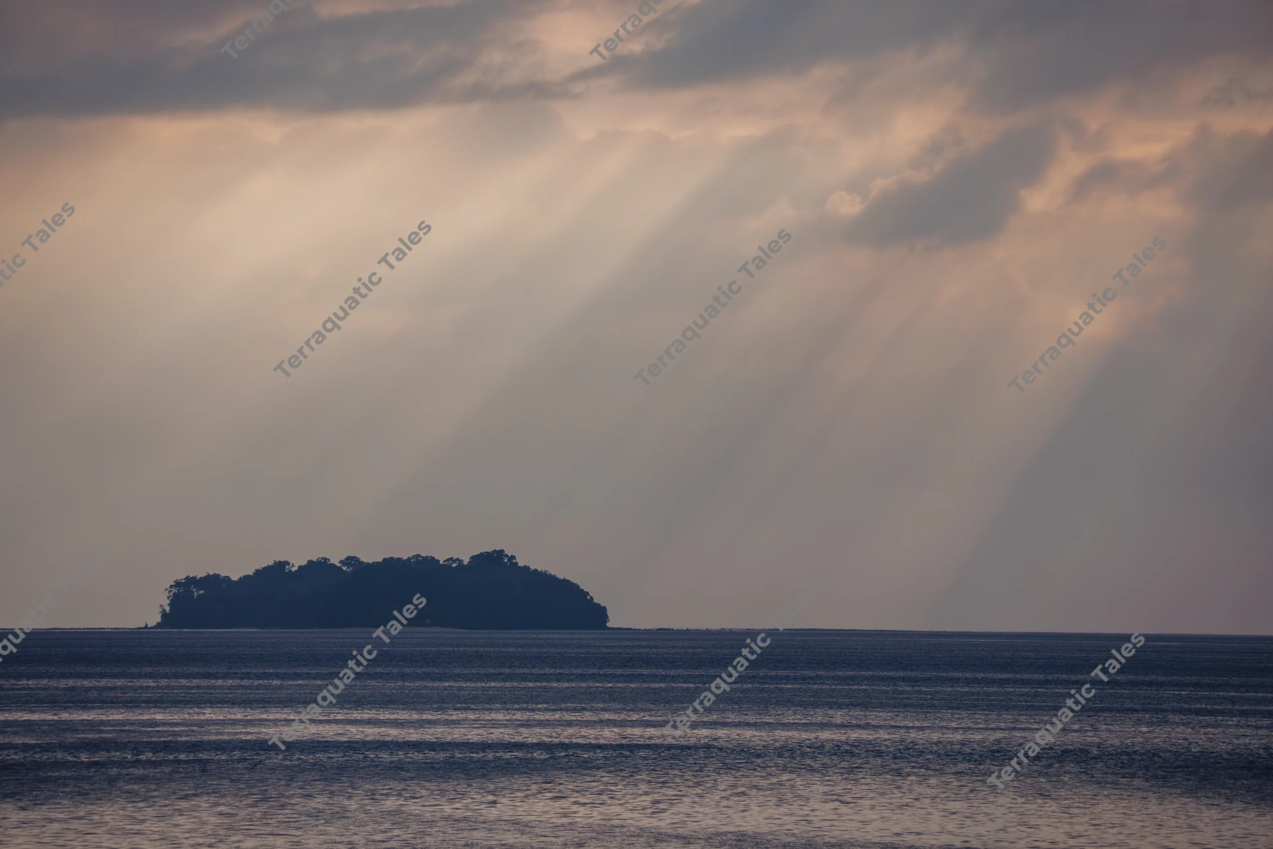 Sunrays beam through clouds above a dark, silhouetted tropical island and calm blue ocean waters in the Andaman Islands.