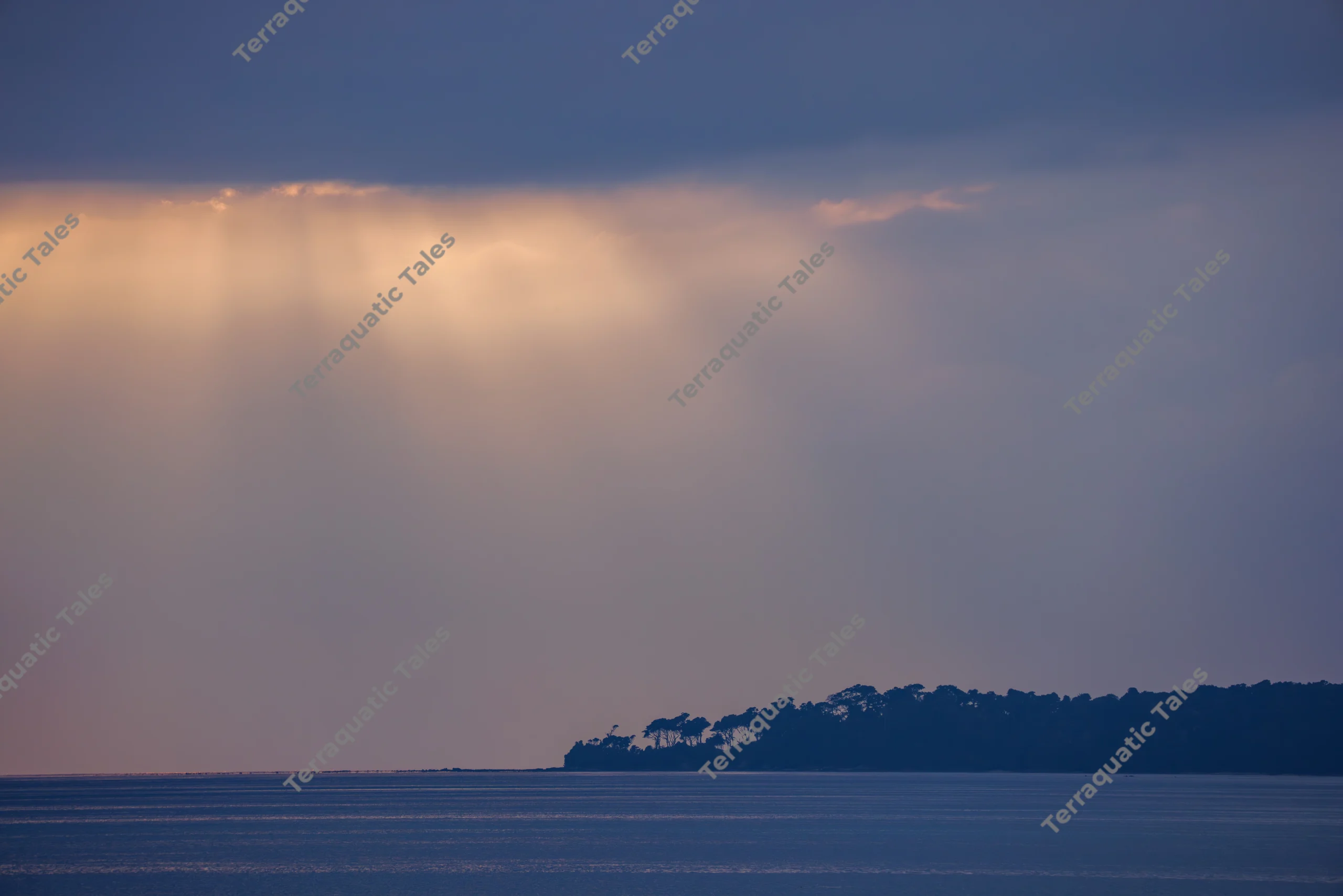 Sunrays beam through clouds above a dark, silhouetted tropical island and calm blue ocean waters in the Andaman Islands.