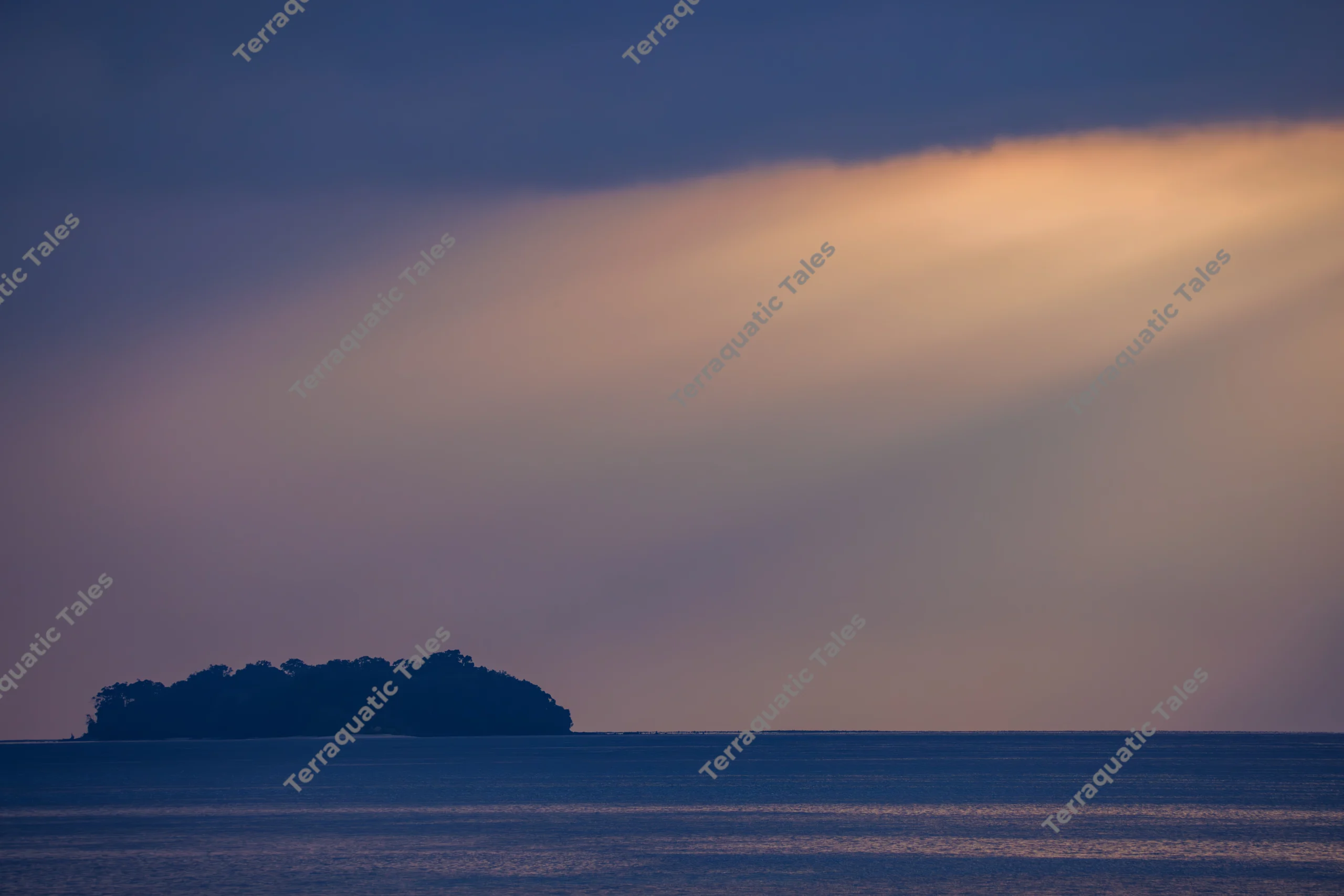 Sunrays beam through clouds above a dark, silhouetted tropical island and calm blue ocean waters in the Andaman Islands.
