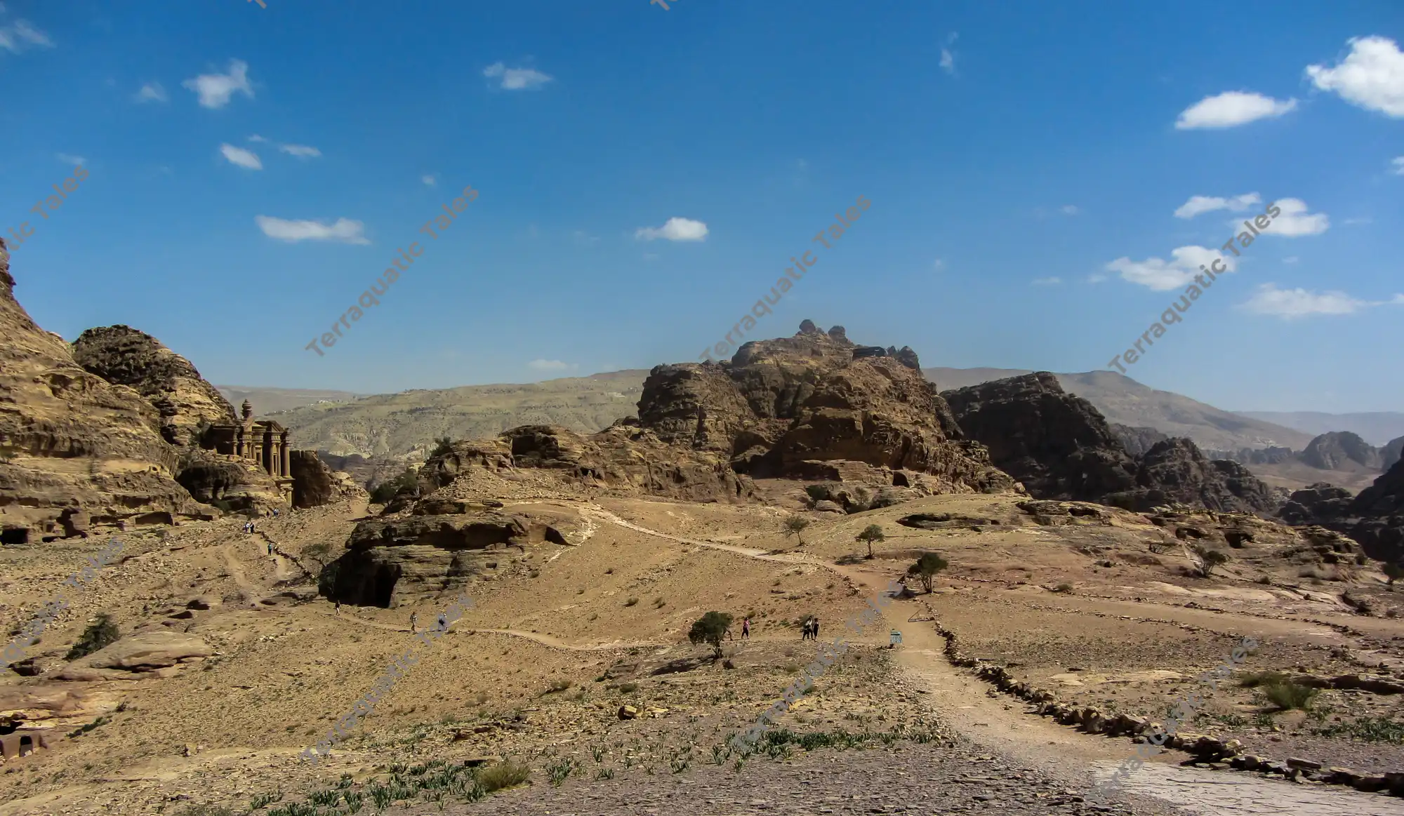 Wide shot of the ancient sandstone facade of Ad Deir, The Monastery, carved into the rocky mountain cliffs of Petra, Jordan, surrounded by desert paths and hills under a clear blue sky.