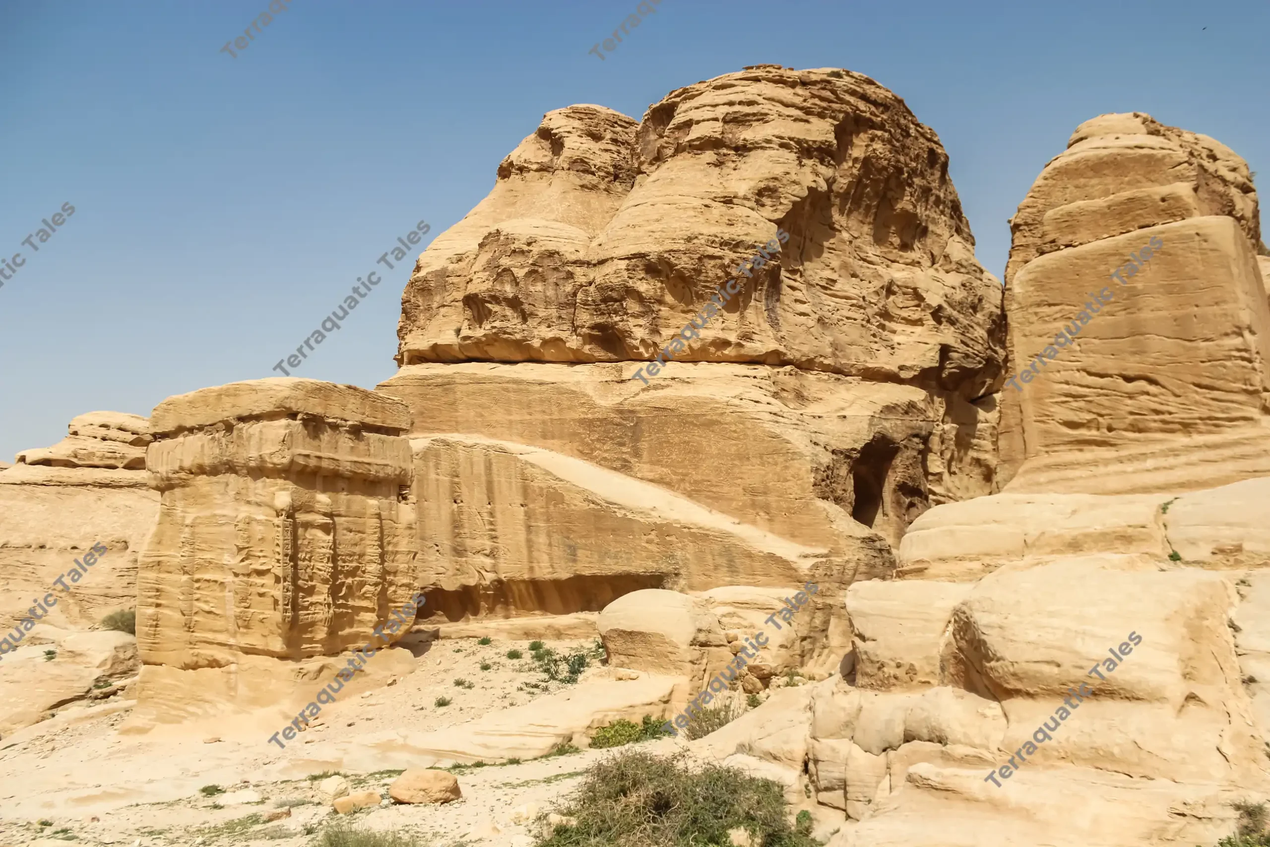 Massive square stone-carved Djinn Blocks made of golden sandstone stand beside weathered desert rock formations under a bright blue sky at Little Petra, Jordan.