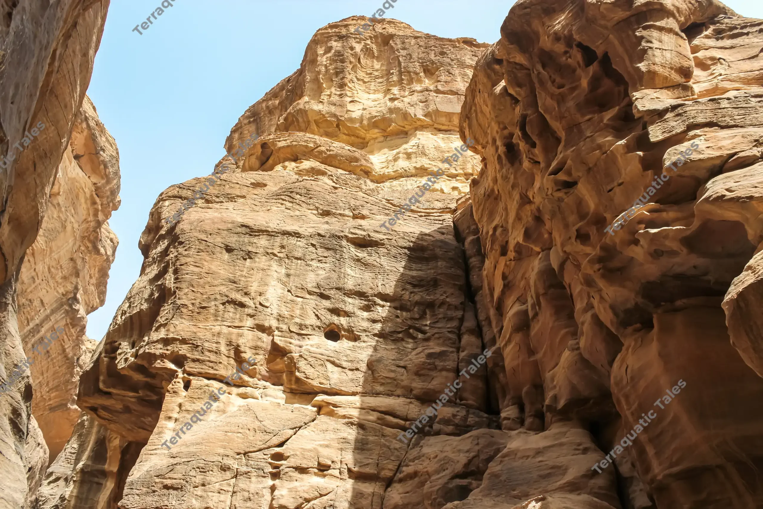 Low-angle view looking straight up at towering, layered golden-brown sandstone cliffs and rock walls framed by a clear blue sky inside Siq al-Barid, Little Petra, Jordan.