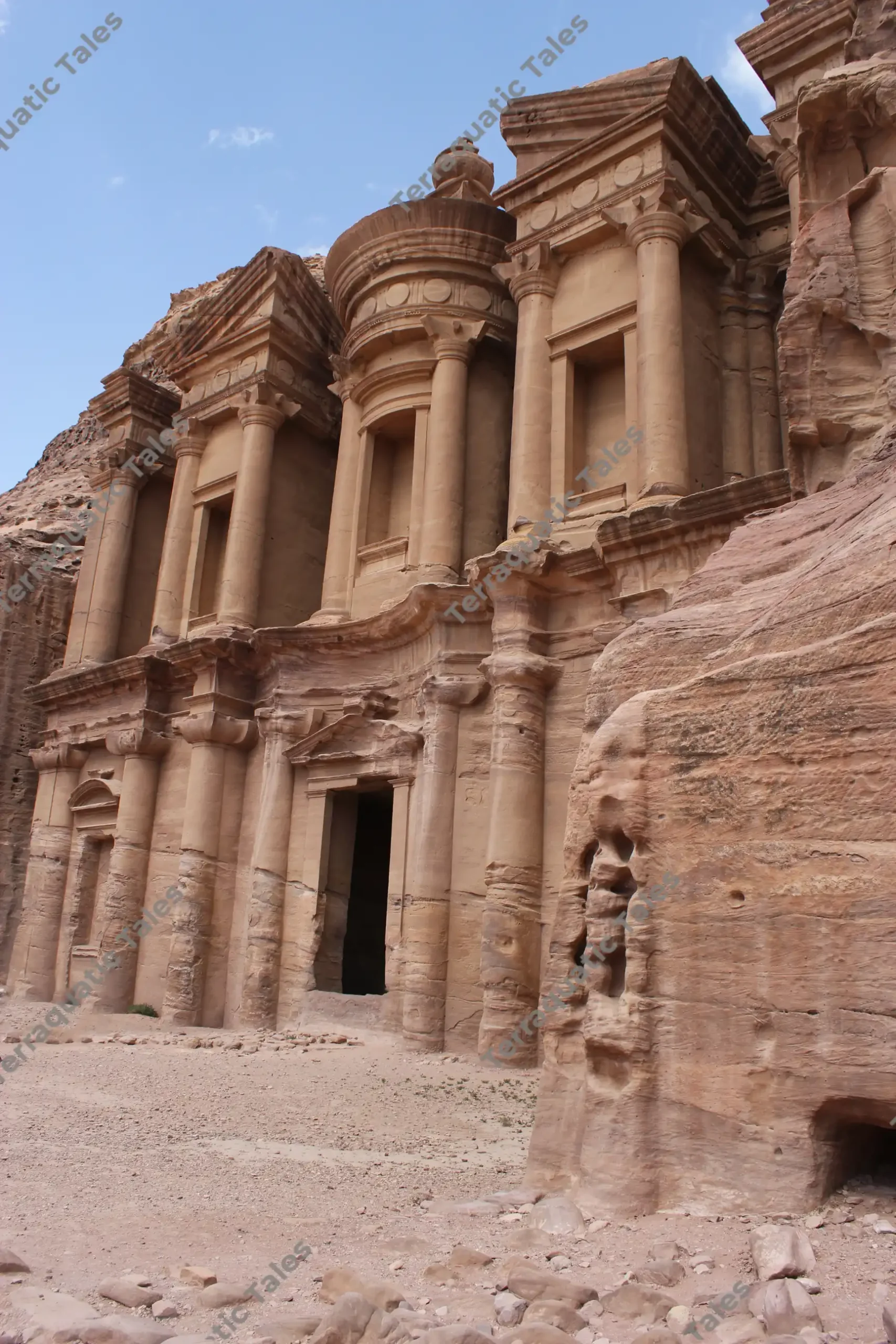A low-angle vertical shot looking up at the grand, sunlit sandstone facade and columns of The Monastery (Ad-Deir) carved into a cliff face in Petra, Jordan.