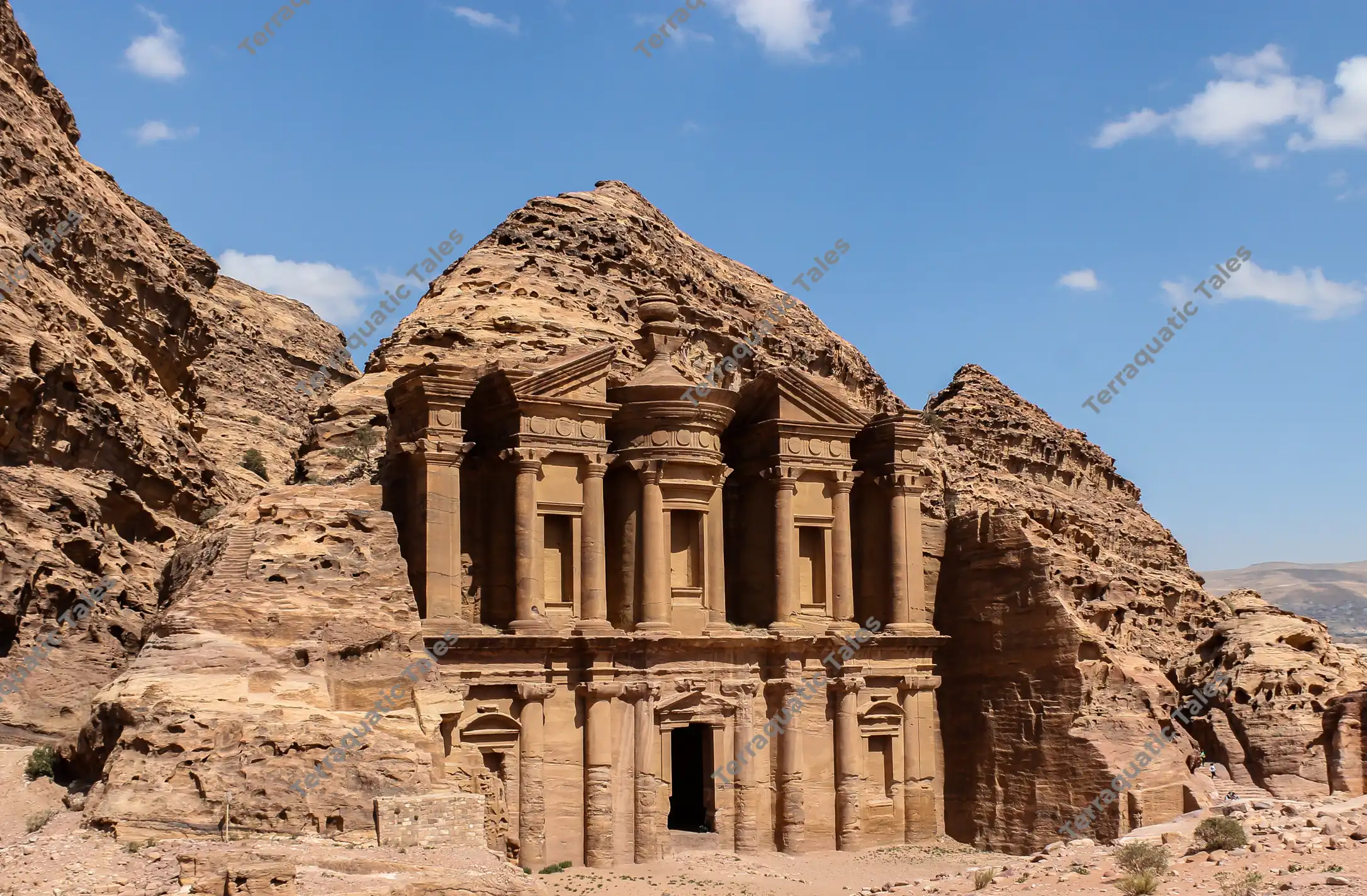 A wide landscape view showing the complete, sunlit golden-tan sandstone facade of The Monastery (Ad-Deir) carved into a rugged mountain peak under a blue sky in Petra, Jordan.