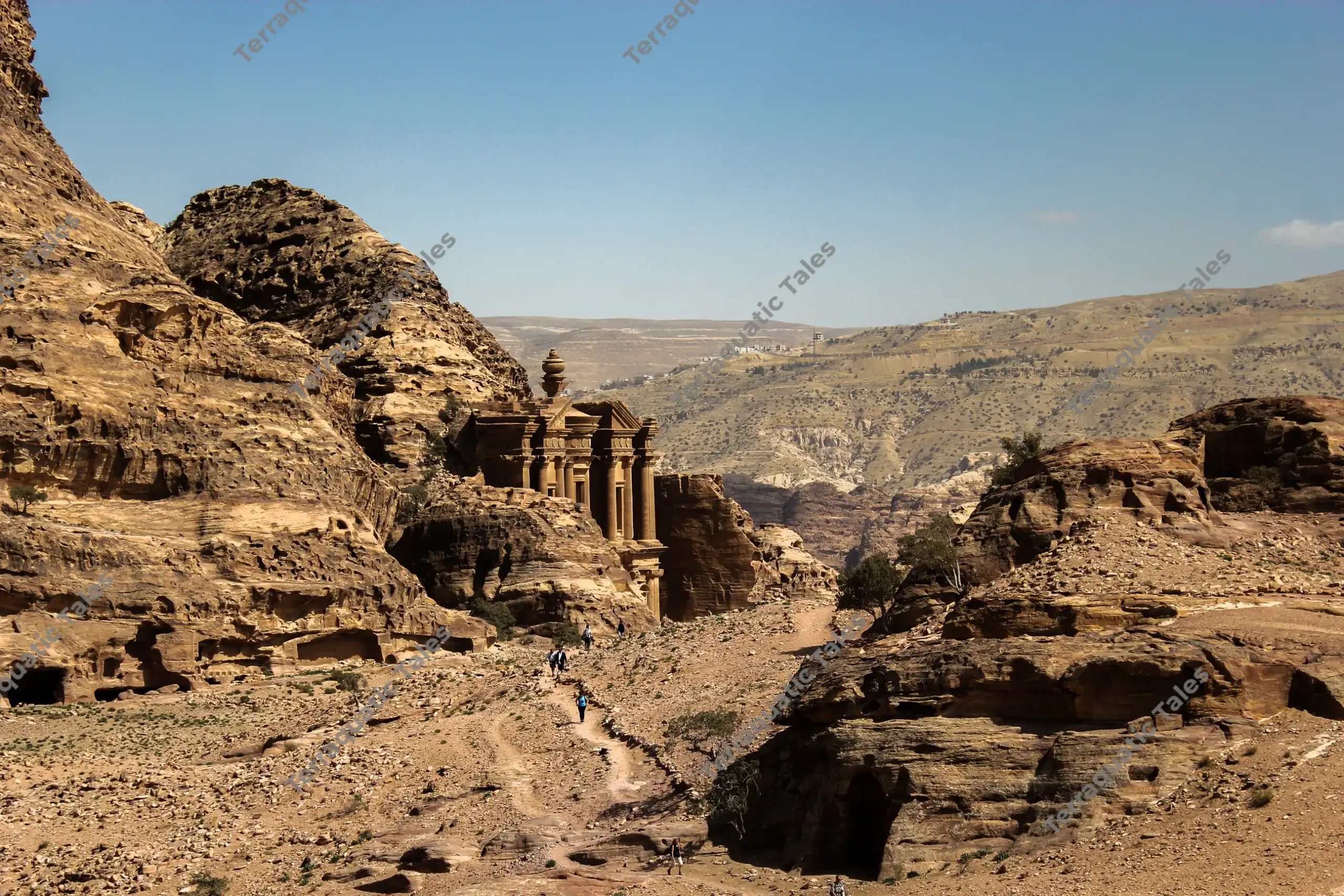 A wide landscape view looking down a desert mountain trail toward the carved facade of The Monastery (Ad-Deir) tucked between sandstone cliffs in Petra, Jordan.