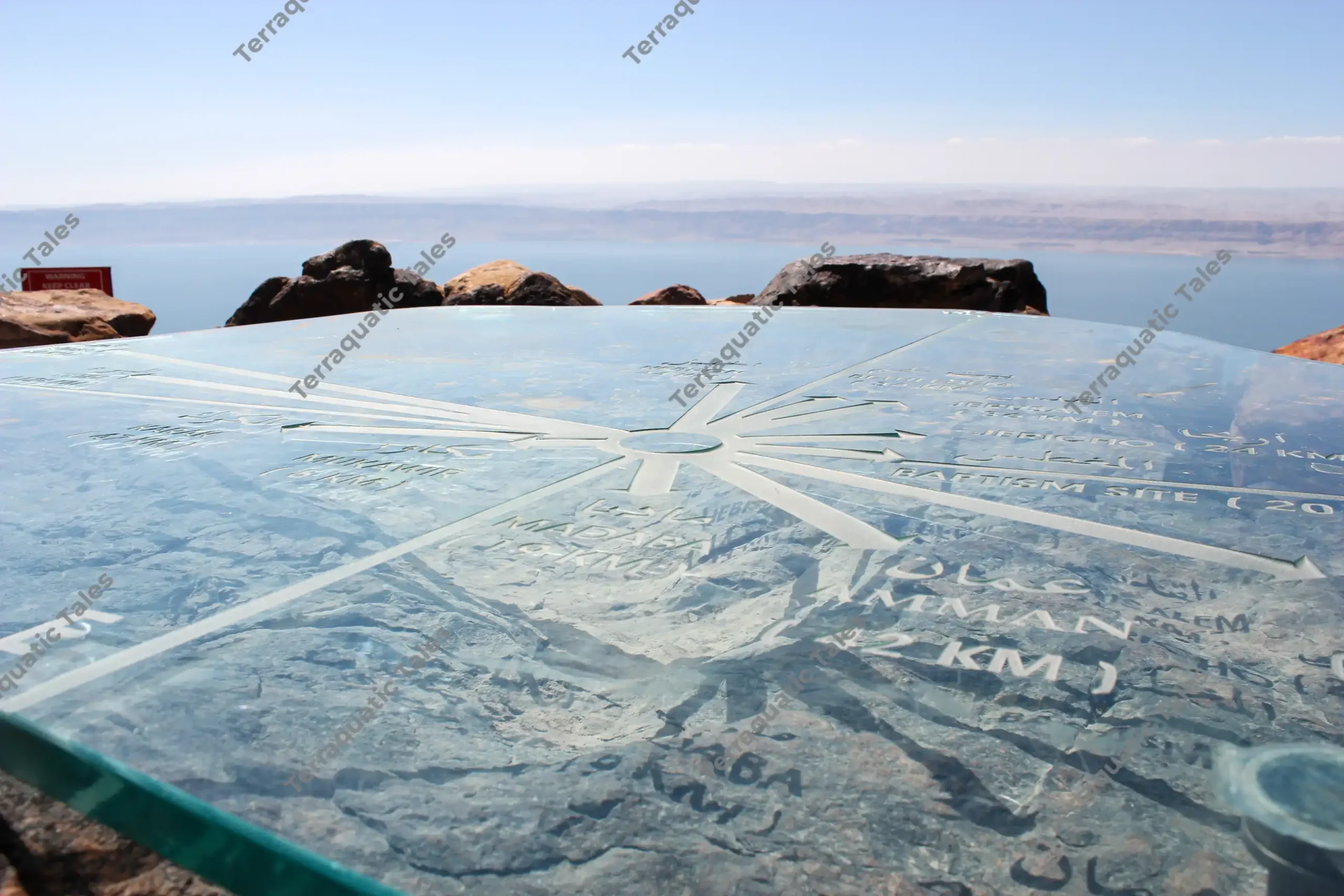 A close-up photograph of an engraved glass orientation map showing directions and distances to regional sites, with the blurry blue Dead Sea and mountains in the background, Jordan.